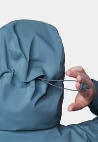 Niebieska kurtka wodoodporna z kapturem, z regulowanym sznurkiem. Zbliżenie na dłoń trzymającą zwężacz do ściągnięcia kaptura.