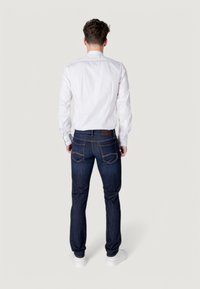 Camisa blanca de manga larga metida dentro de unos vaqueros azul oscuro con una textura suave, que presenta un parche de cuero marrón y costuras visibles en los bolsillos.