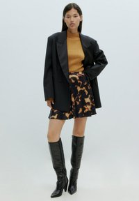 Schwarze, doppelt geknöpfte Blazerjacke über einem senfgelben, gerippten Rollkragenpullover, kombiniert mit einem schwarzen Plisseerock mit orangefarbenem Leopardenmuster und knielangen schwarzen Stiefeln.