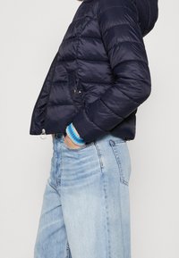 Marinblå puffjacka med huva, quiltat mönster, dragkedja fram och sidofickor. Manchetter med synliga blåvita ränder.