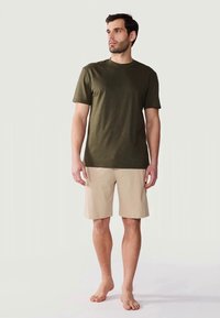 Man standing barefoot wearing an olive green short-sleeve shirt and beige knee-length shorts against a plain light background.