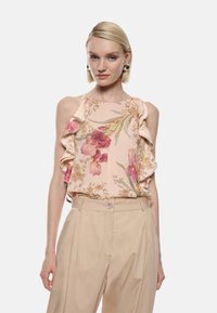 Blusa floreale in morbido tessuto rosa, con spalline arricciate e motivi botanici in rosso e verde, abbinata a pantaloni beige chiaro.