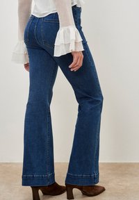 Jeans in denim blu a gamba fluida con vita media, cuciture dettagliate e finitura orlata. Indossati con stivaletti marroni con tacco e una blouse bianca.