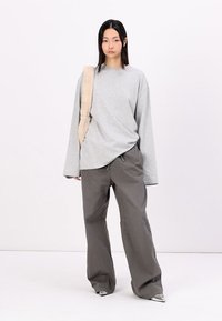Chemise grise à manches longues, à coupe ample, associée à un pantalon large gris ; une sangle légère texturée tombe sur une épaule.