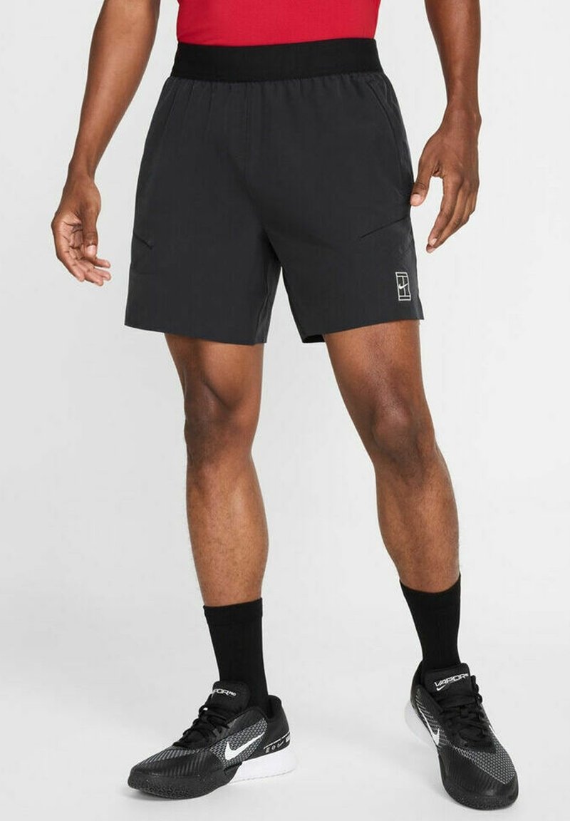 Athlète portant un short sportif noir avec un logo sur la jambe, des chaussettes noires et des baskets Nike noires, debout devant un fond uni.