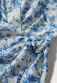 Tissu blanc avec motifs floraux et feuilles bleues, froncé et noué en un nœud au centre, montrant la texture et les plis.