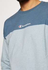 Světle modrý dlouhý rukáv mikiny s tmavě modrým vodorovným pruhem, s bílým logem "Champion" na hrudi. Měkká textura.