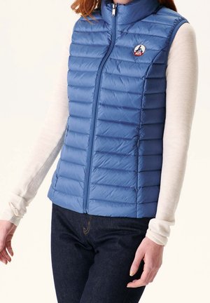 Frau trägt eine hellblaue, gesteppte ärmellose Jacke über einem langärmligen cremefarbenen Shirt und dunklen Jeans, steht mit entspannten Armen.