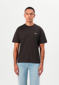 Black cotton t-shirt with a round neckline and short sleeves. Features a small green Lacoste logo on the upper left chest area.