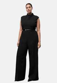 Top negro sin mangas con cuello alto, combinado con pantalones anchos negros. Presenta un cinturón negro delgado en la cintura. Textura de tela suave.