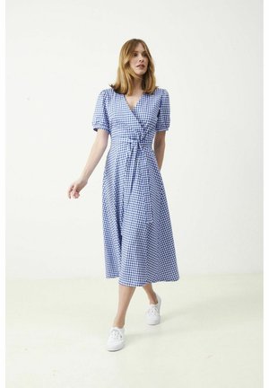 Nainen kävelee yllään sinivalkoruudullinen kietaisumekko, jossa on pussihihat, ja valkoiset tennarit yksivärisellä vaalealla taustalla.