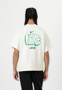 Krēma krāsas kokvilnas t-krekls ar lielu zaļu un melnu grafiku ar uzrakstu "NIKE" aizmugurē, ar apaļu kakla izgriezumu un īsām piedurknēm.