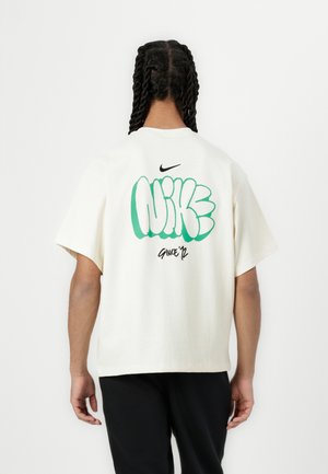 Kremowa bawełniana koszulka z dużą zieloną i czarną grafiką z napisem "NIKE" na plecach, z okrągłym dekoltem i krótkimi rękawami.