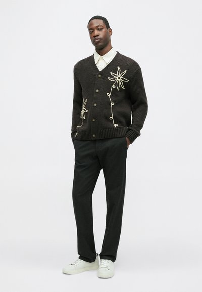 Sötétbarna kötött kardigán fehér virágmintával, V-nyakkal, gombos elülső résszel és bordázott mandzsettával. Sötét nadrággal és fehér sportcipővel párosítva.