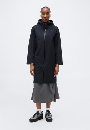 Γυναίκα που φοράει μαύρο αδιάβροχο με κουκούλα πάνω από γκρι midi φούστα και γκρι παπούτσια πεζοπορίας, στέκεται μπροστά σε απλό λευκό φόντο.