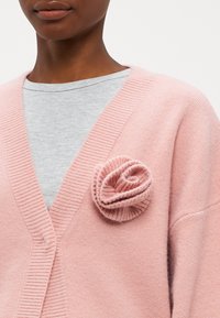 Rozā adīta kardigāna ar V veida kaklu, kuram ir ribota tekstūra un rozēs līdzīga apdare kreisajā pusē. Valkāta virs pelēka topa.