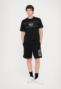 Black cotton t-shirt with white logo and horizontal stripes, paired with black shorts featuring a white logo. Model wears white sneakers.