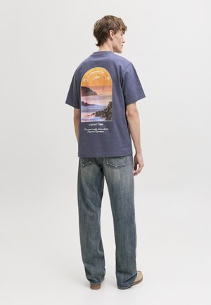 Junger Mann von hinten, trägt ein blaues T-Shirt mit Grafik eines Sonnenuntergangs über dem Ozean und felsiger Küste, kombiniert mit locker sitzenden blauen Jeans und beigen Schuhen.