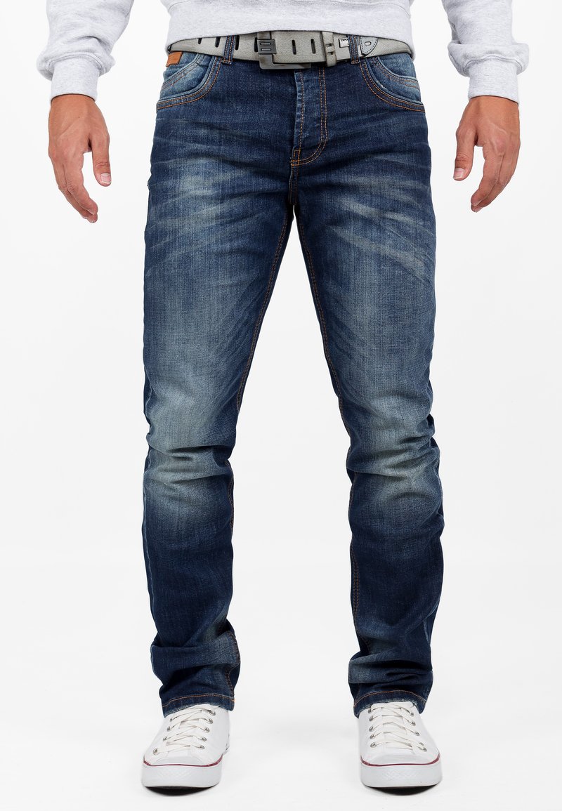 Jeans en denim bleu foncé avec une légère décoloration sur les cuisses, des coutures orange et une ceinture grise. Coupe droite, détails de poches subtils et ourlets retournés.