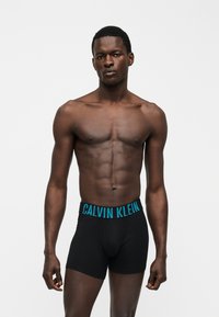 Μυώδης άνδρας με σκούρο δέρμα, φοράει μαύρα boxer Calvin Klein με μπλε λογότυπο, στέκεται μπροστά σε απλό λευκό φόντο.