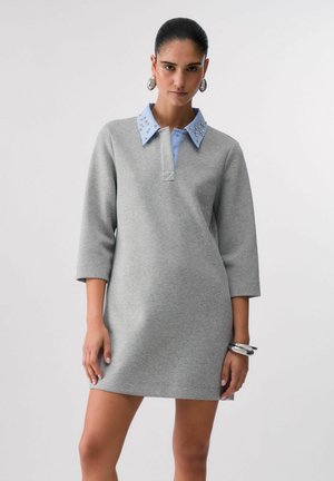 Kobieta w szarej sukience z rękawem do łokcia, z jasnoniebieskim, ozdobionym kołnierzykiem, dużymi srebrnymi kolczykami i srebrnymi bransoletkami na jednym nadgarstku.