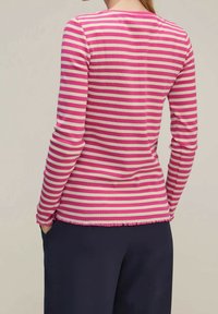 Een persoon in een roze en witte gestreepte langarmshirt met ruches en een donkere broek, staand met de handen in de zakken, van achter bekeken.