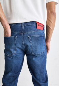 Granatowe spodnie dżinsowe o prostym kroju, z dwoma tylnymi łatkami oraz czerwonym oznaczeniem "HUGO" na tylnej części pasa.