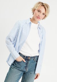 Chemise boutonnée à rayures bleues et blanches avec des manches longues, fabriquée en tissu léger, portée ouverte sur un t-shirt blanc et un jean en denim.