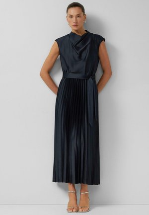 Kobieta w ciemnogranatowej, plisowanej sukience midi bez rękawów, z marszczonym dekoltem i paseczkiem w talii, stoi na tle jasnoszarej ściany.