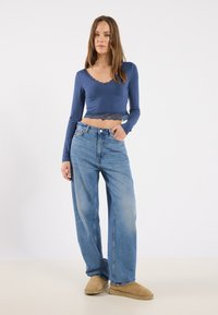 Tettfitt, langermet blå crop topp med blondekant, kombinert med høyt liv lyseblå vide jeans og beige slip-on sko.