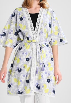 Kobieta w kwiecistym kardiganie w stylu kimono z żółtymi, czarnymi i lawendowymi kwiatami, koronkowym wykończeniem oraz wiązanym paskiem w talii z chwostami.