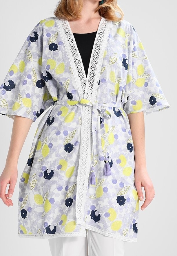 Femme portant un cardigan style kimono à fleurs jaunes, noires et lavande, avec bordure en dentelle et ceinture nouée à la taille avec des pompons.