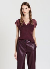 Giovane donna che indossa un top in pizzo bordeaux a maniche corte e pantaloni in pelle di un viola scuro a vita alta, in piedi contro uno sfondo semplice.