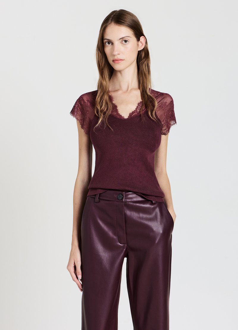 Giovane donna che indossa un top in pizzo bordeaux a maniche corte e pantaloni in pelle di un viola scuro a vita alta, in piedi contro uno sfondo semplice.