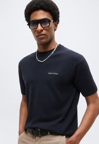 Άνδρας που φοράει μαύρο πουκάμισο Marc O'Polo, αλυσίδα λαιμού, μαύρα γυαλιά ηλίου και μπεζ παντελόνι με μαύρη ζώνη, στέκεται μπροστά σε απλό φόντο.