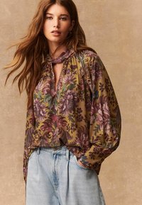 Blusa con motivo floreale in verde oliva, con maniche lunghe, scollatura a chiave e tessuto drappeggiato, abbinata a jeans chiari a vita alta.
