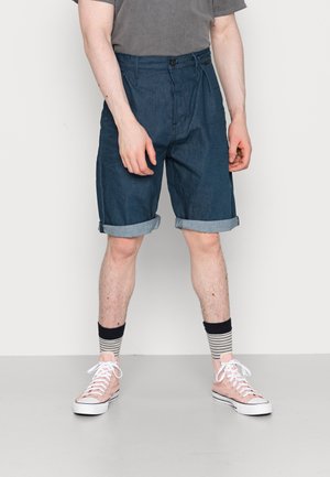 Person iført opkørte mørkeblå denimshorts, grå t-shirt, stribede sorte og hvide sokker og lyserøde høje sneakers.