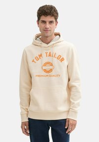 Beige Kapuzenpullover mit Kapuze und frontaler Tasche, mit orangefarbener Schrift "TOM TAILOR PREMIUM QUALITY" und einem runden Logodetail. Sanfte Textur.