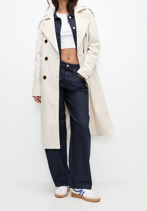 Beiger Trenchcoat mit schwarzen Knöpfen, getragen über einem weißen, gekürzten Oberteil und blauen, Nadelstreifenhosen. Weiße Sneaker mit blauen Streifen.