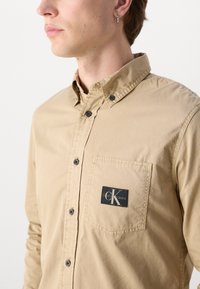 Camicia button-up color beige con colletto, dotata di taschino sul petto e una toppa nera con logo. I dettagli includono bottoni neri e una texture liscia.