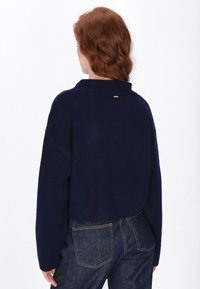 Pulover tricotat scurt, de culoare bleumarin, cu guler, având umerii lăsați și un mic detaliu metallic pe spate. Asortat cu blugi închis la culoare.