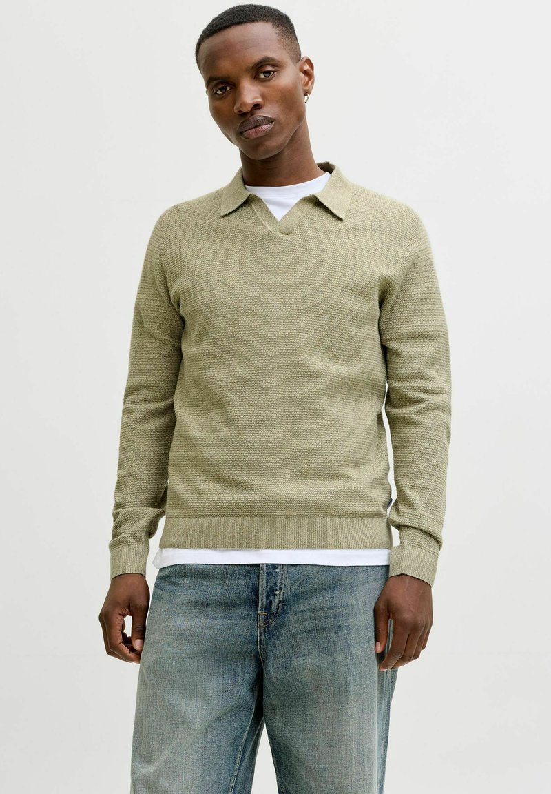 Pull en tricot vert clair avec col en V et col, tissu texturé, poignets côtelés, porté sur une chemise blanche, associé à un jean bleu.
