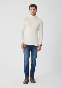 Beiger Rollkragenpullover, figurbetont, mit langen Ärmeln; kombiniert mit blauen Denim-Jeans und braunen Stiefeletten. Einfache und neutrale Farbpalette.
