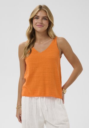 Femeie cu păr lung și blond, purtând un top portocaliu tricotat fără mâneci și pantaloni albi cu textură, stând cu o mână pe șold.