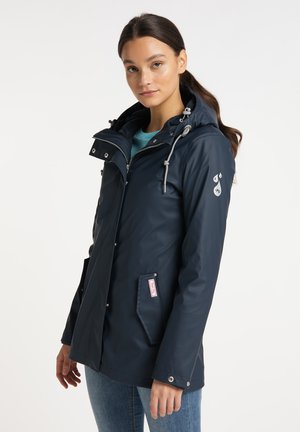 Jonge vrouw met donker haar draagt een marineblauwe regenjas met capuchon over een blauwgroen shirt en blauwe spijkerbroek, staand tegen een effen lichte achtergrond.