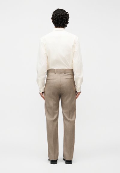 Άνδρας με σγουρά σκούρα μαλλιά που φοράει κρεμ πουκάμισο με μακριά μανίκια και μπεζ παντελόνι υψηλής μέσης με ρίγες, στέκεται με την πλάτη προς τα έξω πάνω σε απλό φόντο.