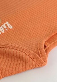 Tissu orange à côtes avec une texture douce, présentant du texte blanc. Le bord du vêtement est terminé avec un ourlet et de jolis détails de couture.