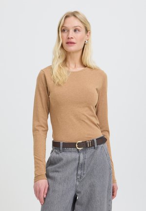 Femme blonde portant une chemise beige à manches longues rentrée dans un jean gris taille haute avec une ceinture marron foncé, regardant légèrement sur le côté.