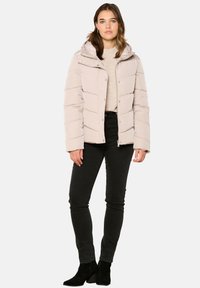 Doudoune de couleur beige clair, dotée d'une capuche, de boutons-pression à l'avant et d'un motif matelassé horizontal. Portée par-dessus un pull beige et un pantalon noir.