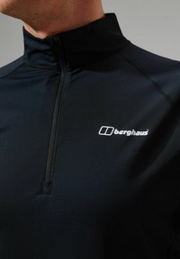 Zwarte quarter-zip pullover van rekbaar materiaal, met een hoge kraag en een klein wit logo op de borst. Soepele textuur.